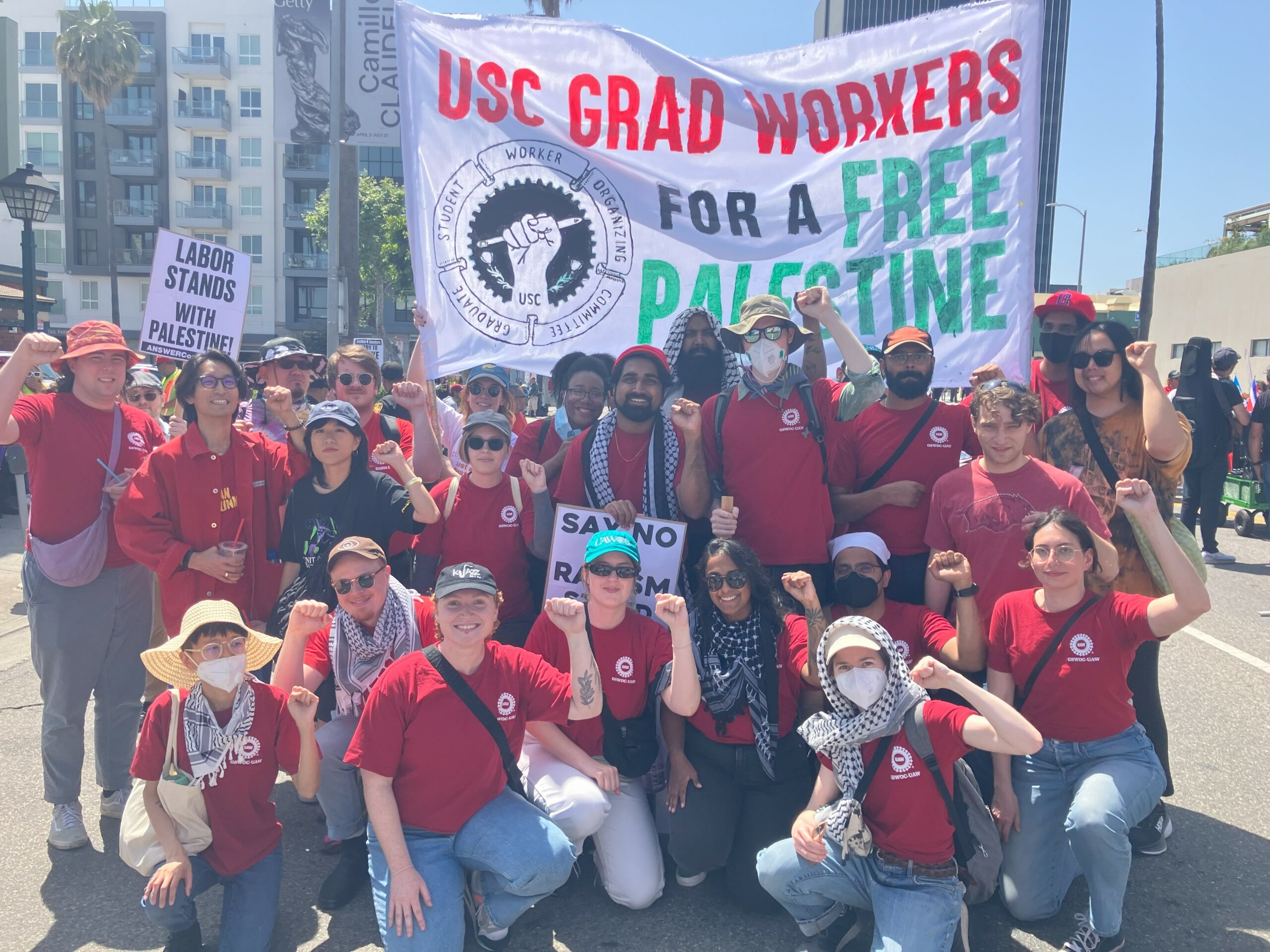 About 25 union members wearing union shirts and keffiyehs raising their fists in front of a banner reading "USC Grad Workers for a Free Palestine" at the May Day March