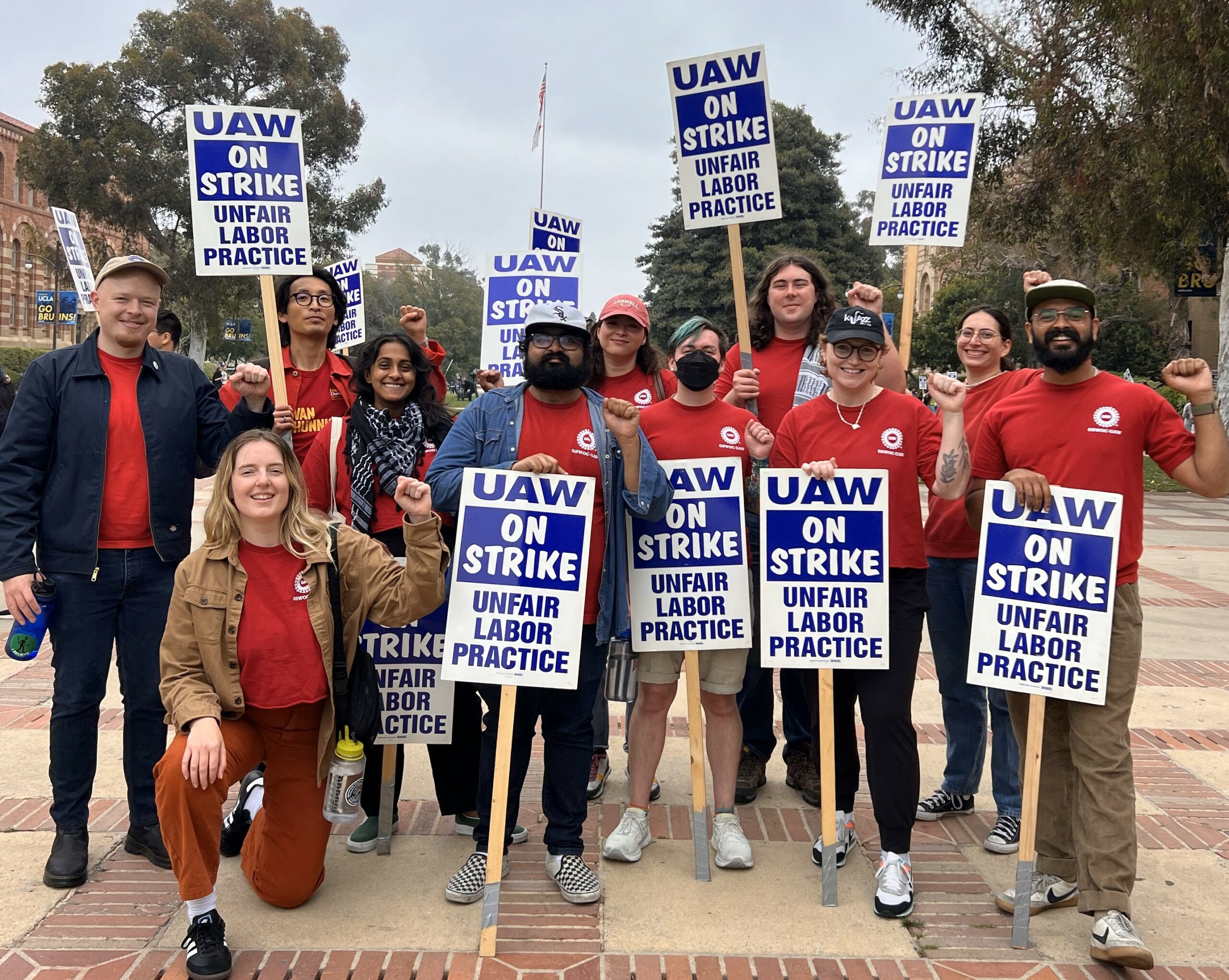 11 union members holding "UAW On Strike: Unfair Labor Practice" picket signs and raising their fists